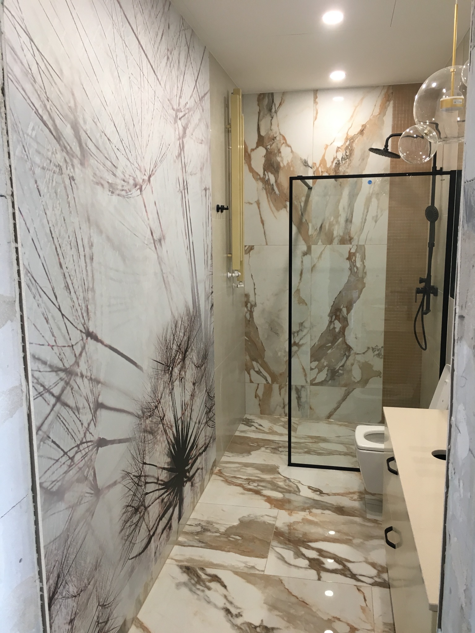 Nowoczesna łazienka z marmurowymi płytkami w odcieniach bieli i brązu, kabiną prysznicową w czarnej ramie, umywalką z szafką i toaletą. Na ścianie dekoracyjny wzór dmuchawca.
