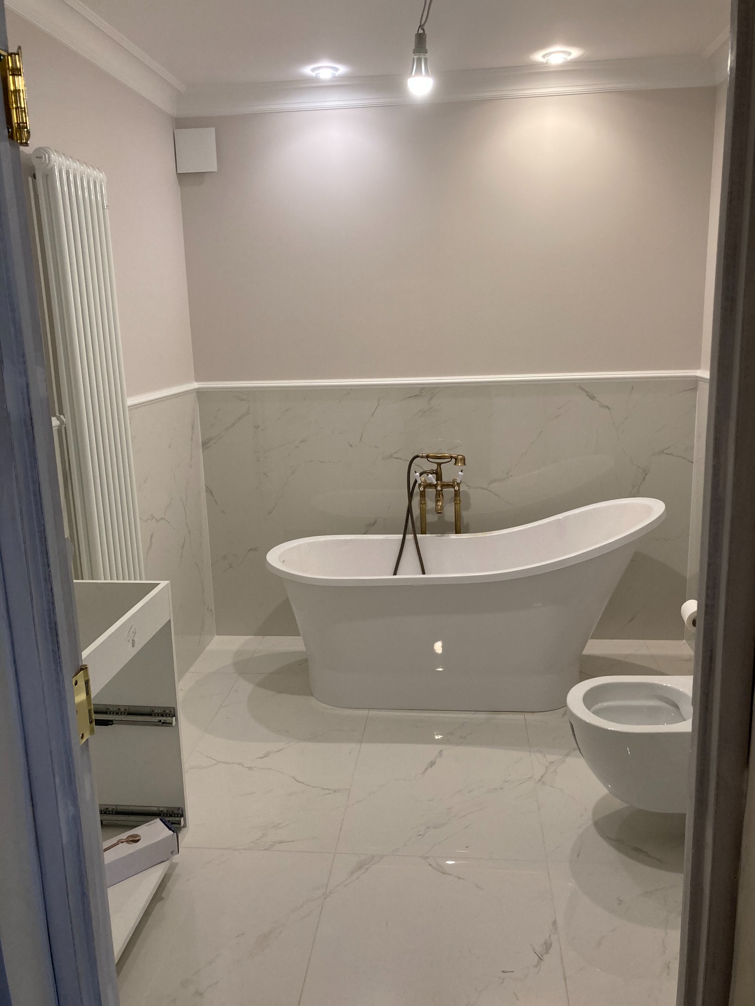 Nowoczesna łazienka z wolnostojącą białą wanną z złotą armaturą, podwieszaną toaletą i marmurowymi płytkami, widok na biały grzejnik i częściowo zmontowaną szafkę pod umywalkę.