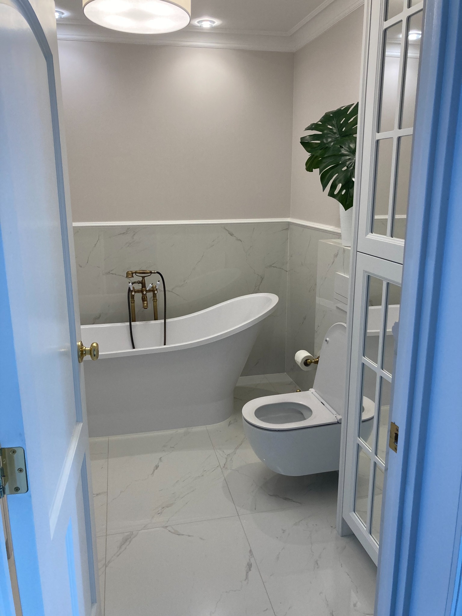 Elegancka łazienka w odcieniach bieli i szarości z wolnostojącą wanną na złotych nóżkach, toaletą typu rimless oraz dużą rośliną doniczkową w tle, widoczna przez uchylone drzwi.