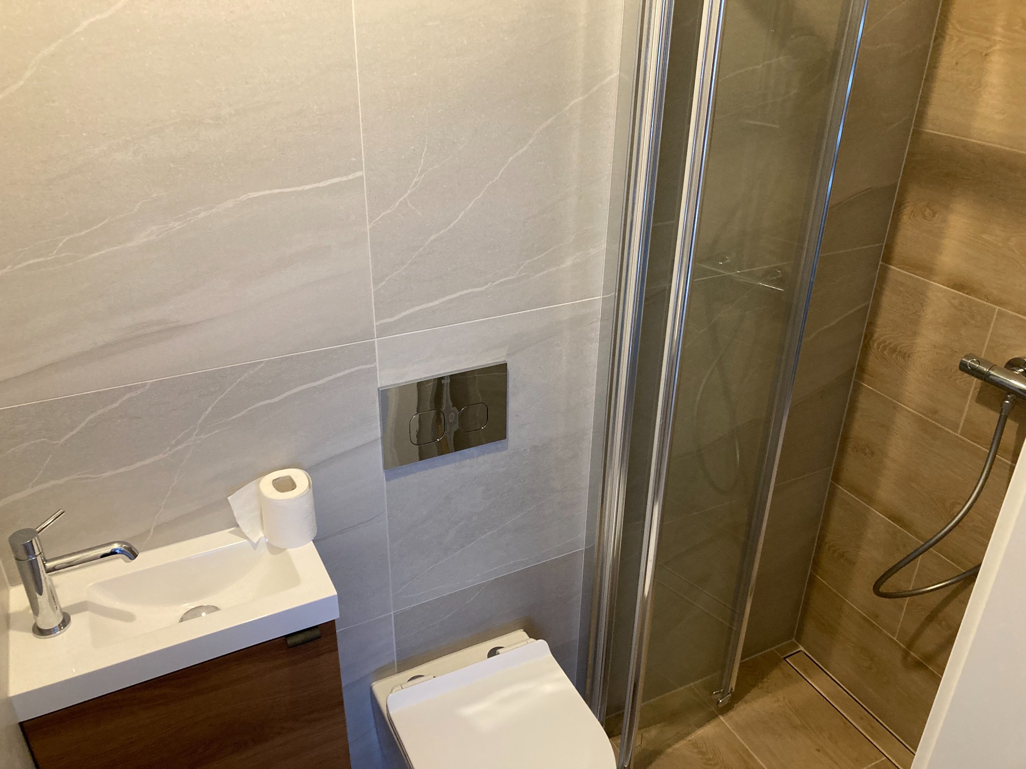 Nowoczesna łazienka z szarymi płytkami imitującymi marmur, wiszącą umywalką z brązową szafką, toaletą i kabiną prysznicową z odpływem liniowym.