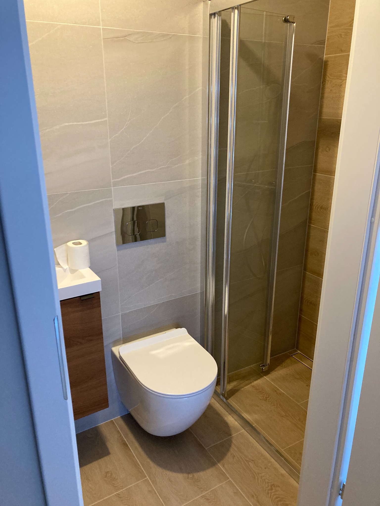 Nowoczesna łazienka z beżowymi płytkami imitującymi marmur, wisząca toaleta, kabina prysznicowa ze składanymi drzwiami i brązowa szafka pod umywalką z rolką papieru toaletowego.