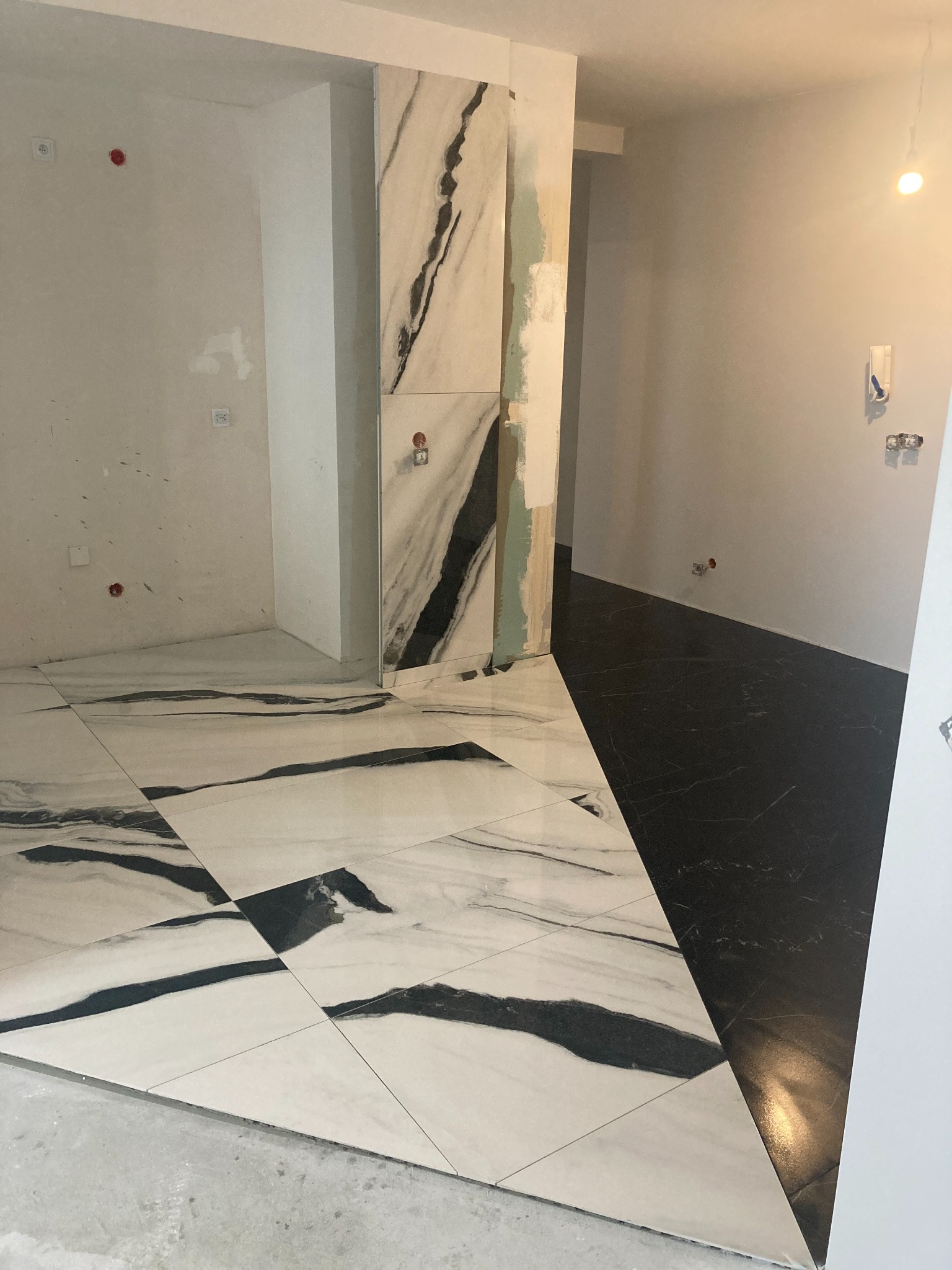 Łazienka w trakcie prac wykończeniowych; podłoga wyłożona białymi i czarnymi płytkami imitującymi marmur, ściany surowe, widoczne instalacje elektryczne i hydrauliczne.