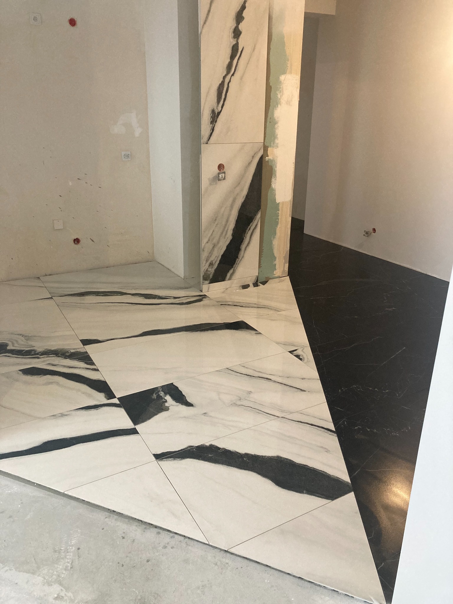 Układanie płytek podłogowych w łazience, widoczne białe płytki z czarnymi żyłami oraz czarne płytki, surowe ściany z widocznymi puszkami elektrycznymi.