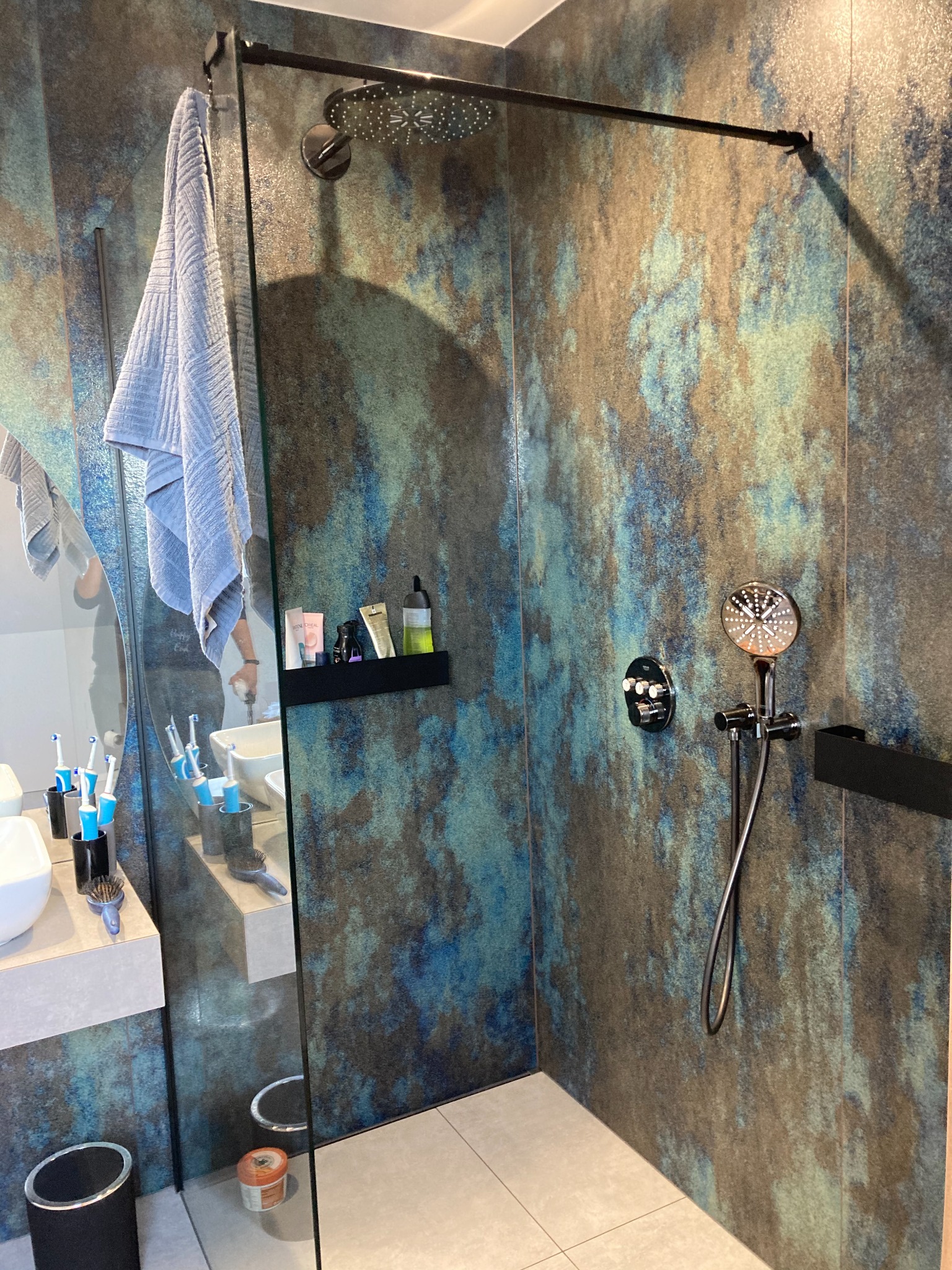 Nowoczesna kabina prysznicowa walk-in z deszczownicą i baterią termostatyczną na ścianie o teksturze imitującej kamień w odcieniach niebieskiego i brązu, z półką na kosmetyki i wiszącym ręcznikiem.