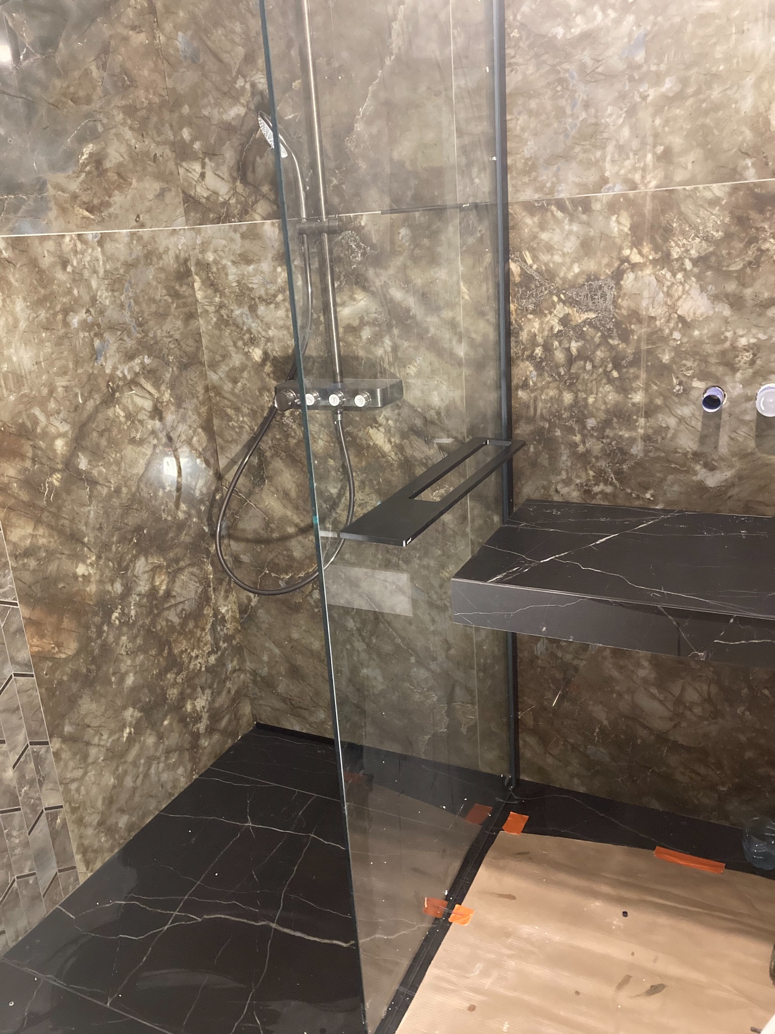 Wykończona łazienka z kabiną prysznicową typu walk-in, brązowe marmurowe płytki na ścianach, czarna marmurowa podłoga i półka, widoczne elementy instalacji prysznicowej