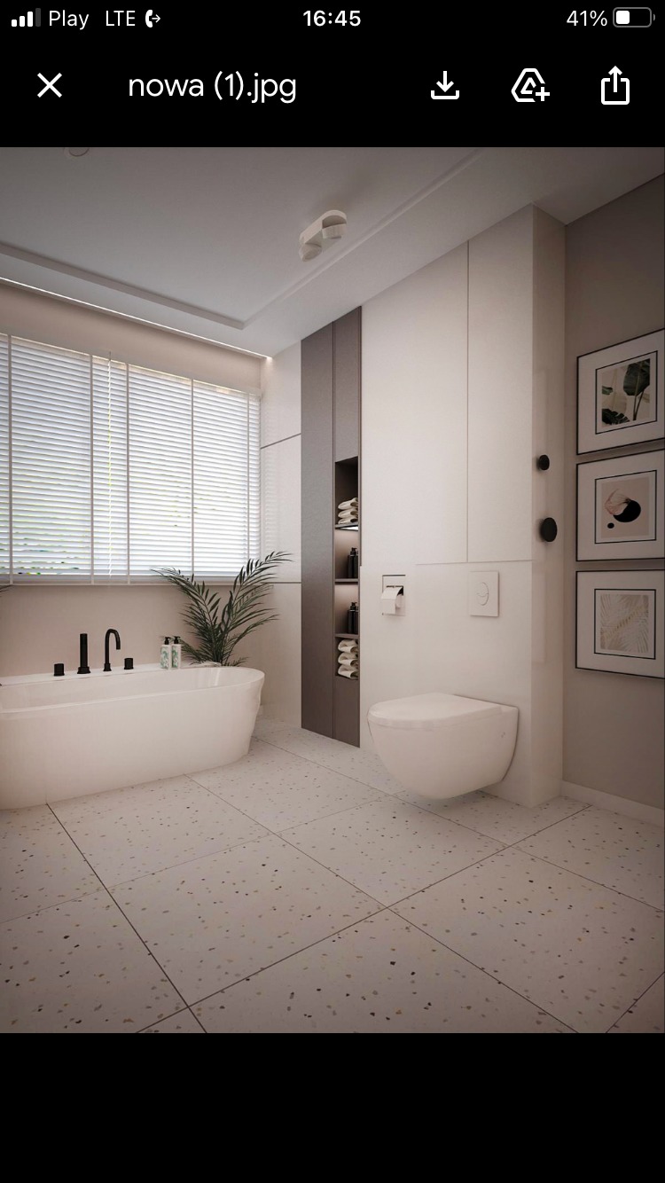 Nowoczesna łazienka z wolnostojącą wanną, wiszącą toaletą, szafką w zabudowie i jasną terakotą z drobnymi plamkami, doświetlona światłem dziennym przez żaluzje.