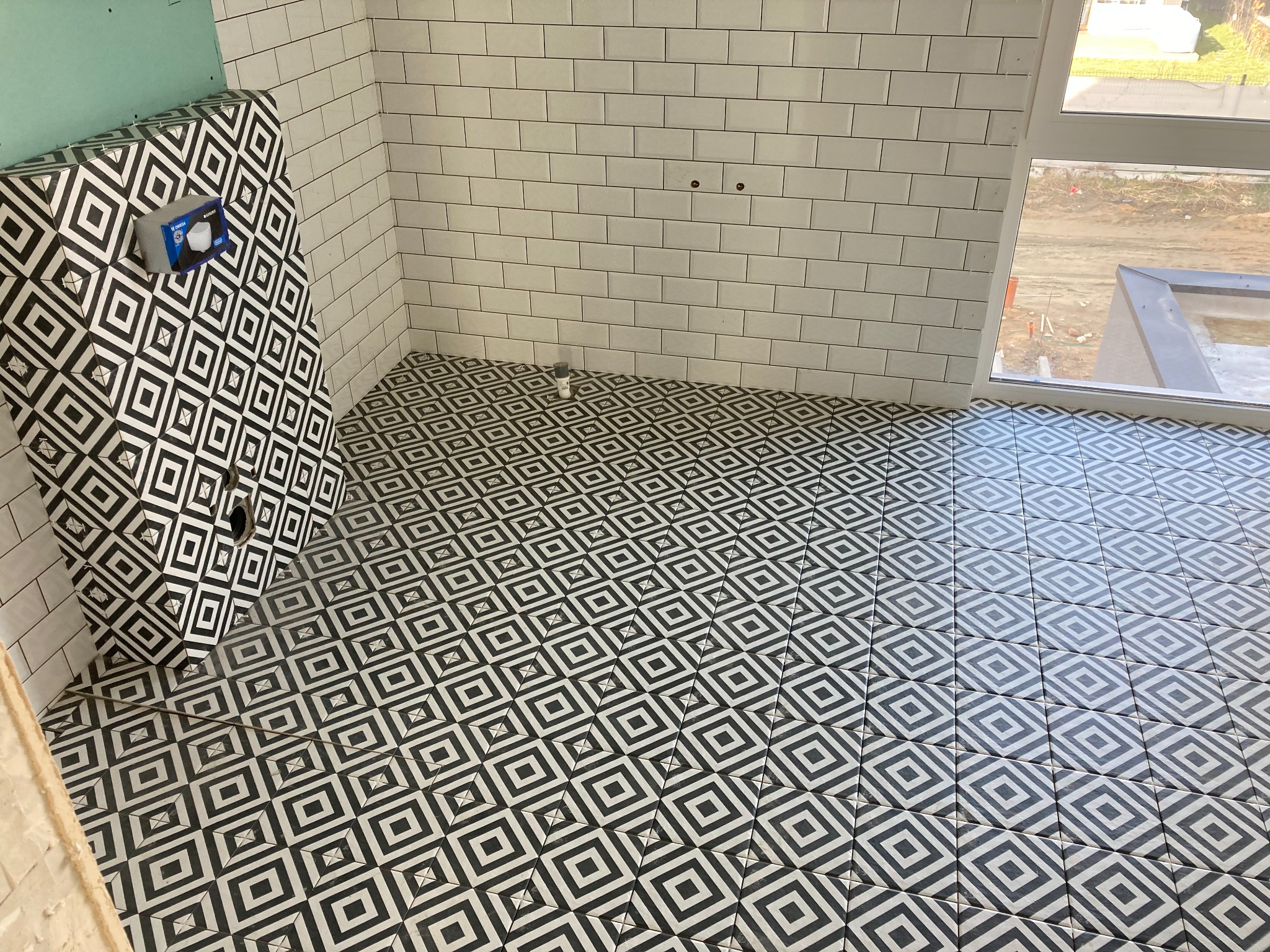 Nowoczesna łazienka w trakcie prac wykończeniowych, z geometryczną czarno-białą posadzką i obudową stelaża podtynkowego, białe płytki na ścianach, okno z widokiem na plac budowy.