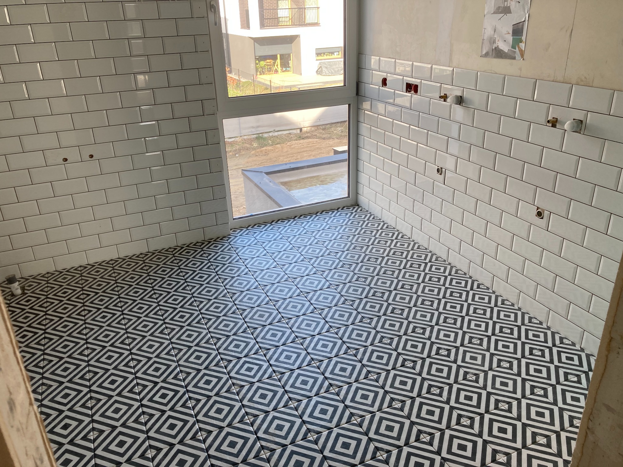 Łazienka w trakcie prac: białe, prostokątne płytki na ścianach ułożone do połowy wysokości, podłoga wyłożona czarno-białą, geometryczną mozaiką, widoczne otwory na instalacje wodne i elektryczne.