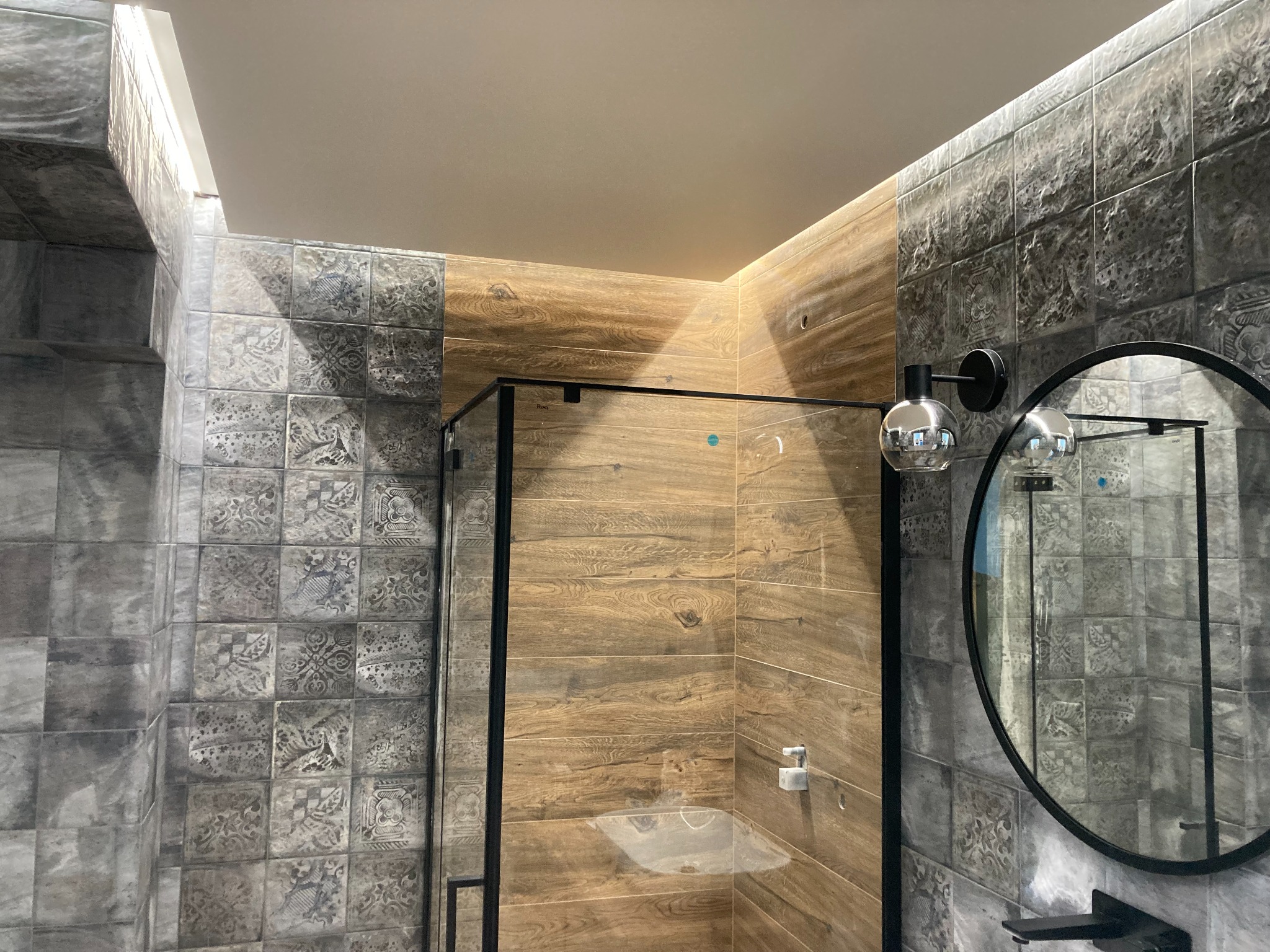 Nowoczesna kabina prysznicowa z drewnopodobnymi płytkami, oświetleniem LED i okrągłym lustrem z czarną ramą w łazience wykończonej szaro-wzorzystymi płytkami.