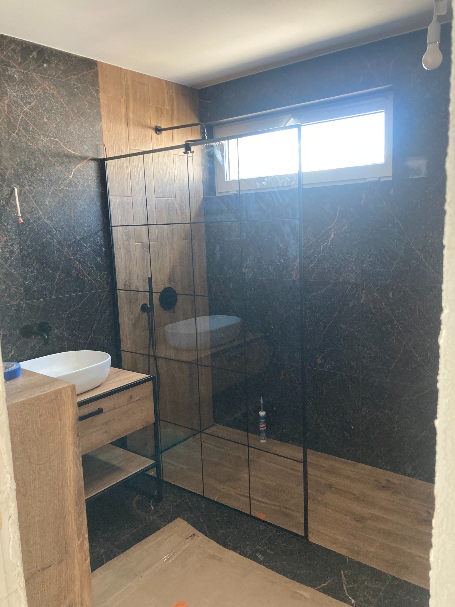 Nowoczesna łazienka w trakcie wykańczania: kabina prysznicowa z czarnymi profilami, umywalka nablatowa na drewnianej szafce, ściany wyłożone płytkami imitującymi marmur i drewno