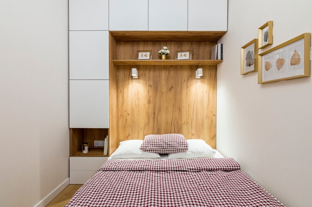 Mała sypialnia z zabudową meblową: białe szafki, drewniany panel za łóżkiem z półkami, oświetlenie punktowe i dekoracje ścienne.