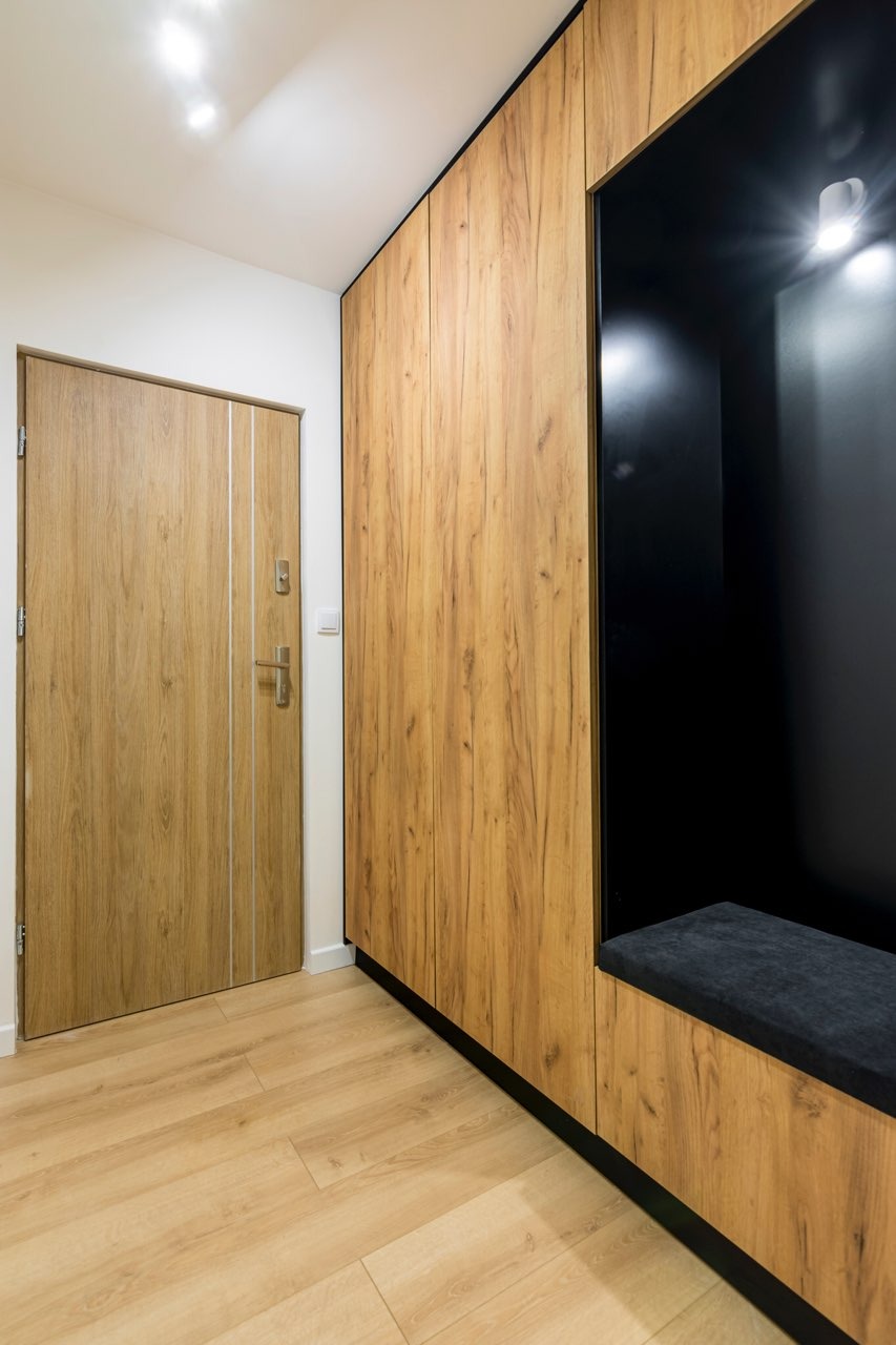 Wnętrze przedpokoju z drewnianą zabudową szafy wnękowej z czarną wnęką na siedzisko i drewnianymi drzwiami wejściowymi z srebrnymi wstawkami.