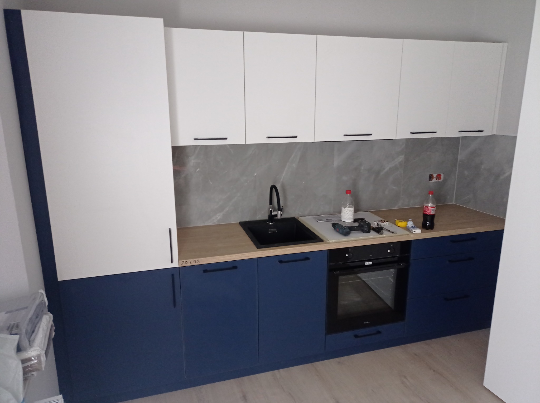 Nowoczesna zabudowa kuchenna w kolorach białym i granatowym, z czarnym zlewem i sprzętem AGD, widoczne narzędzia i napoje na blacie, marmurowa ściana w tle.