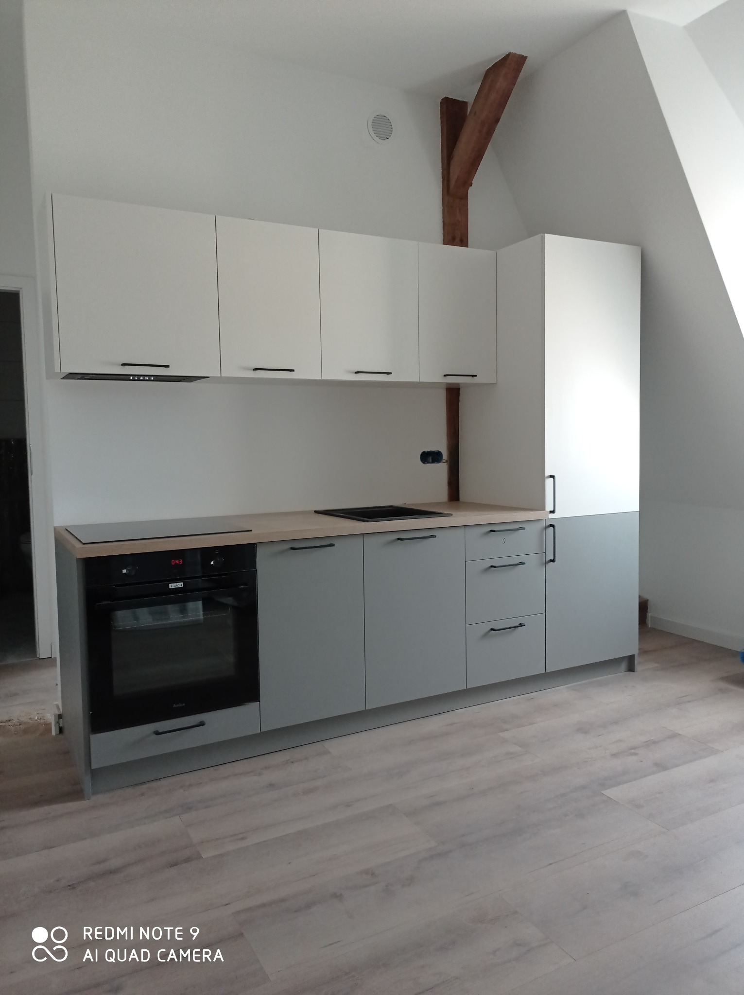 Nowoczesna kuchnia w odcieniach szarości i bieli, z drewnianym blatem i widoczną drewnianą belką stropową, umieszczona na poddaszu ze skośnym sufitem.