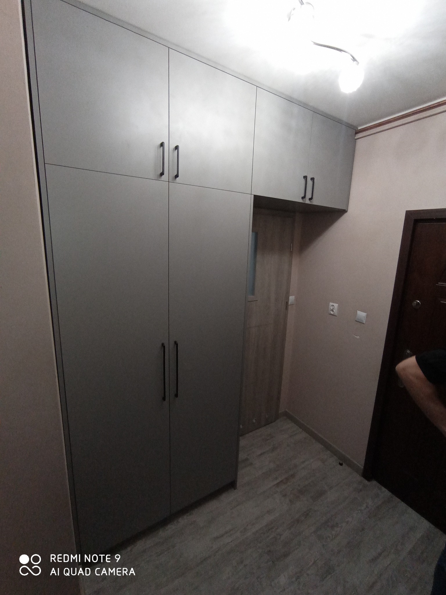 Szara szafa wnękowa z czarnymi uchwytami, zabudowana do sufitu w przedpokoju z drewnopodobną podłogą i widocznymi drzwiami.