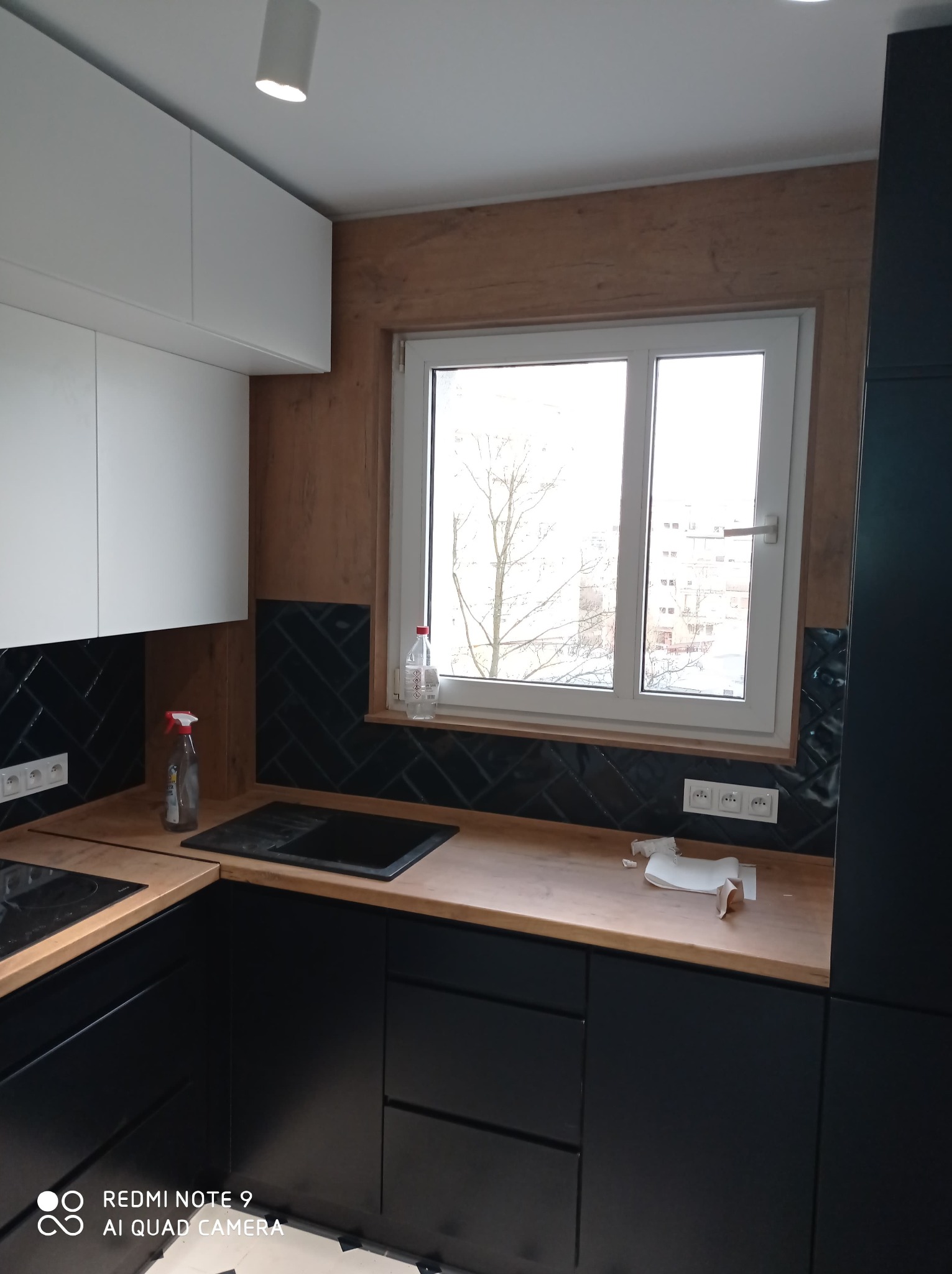 Częściowy widok nowoczesnej kuchni z czarnymi szafkami dolnymi i białymi górnymi, drewnianym blatem, czarnymi płytkami ułożonymi w jodełkę nad blatem, oknem z widokiem na budynki i drzewa,...