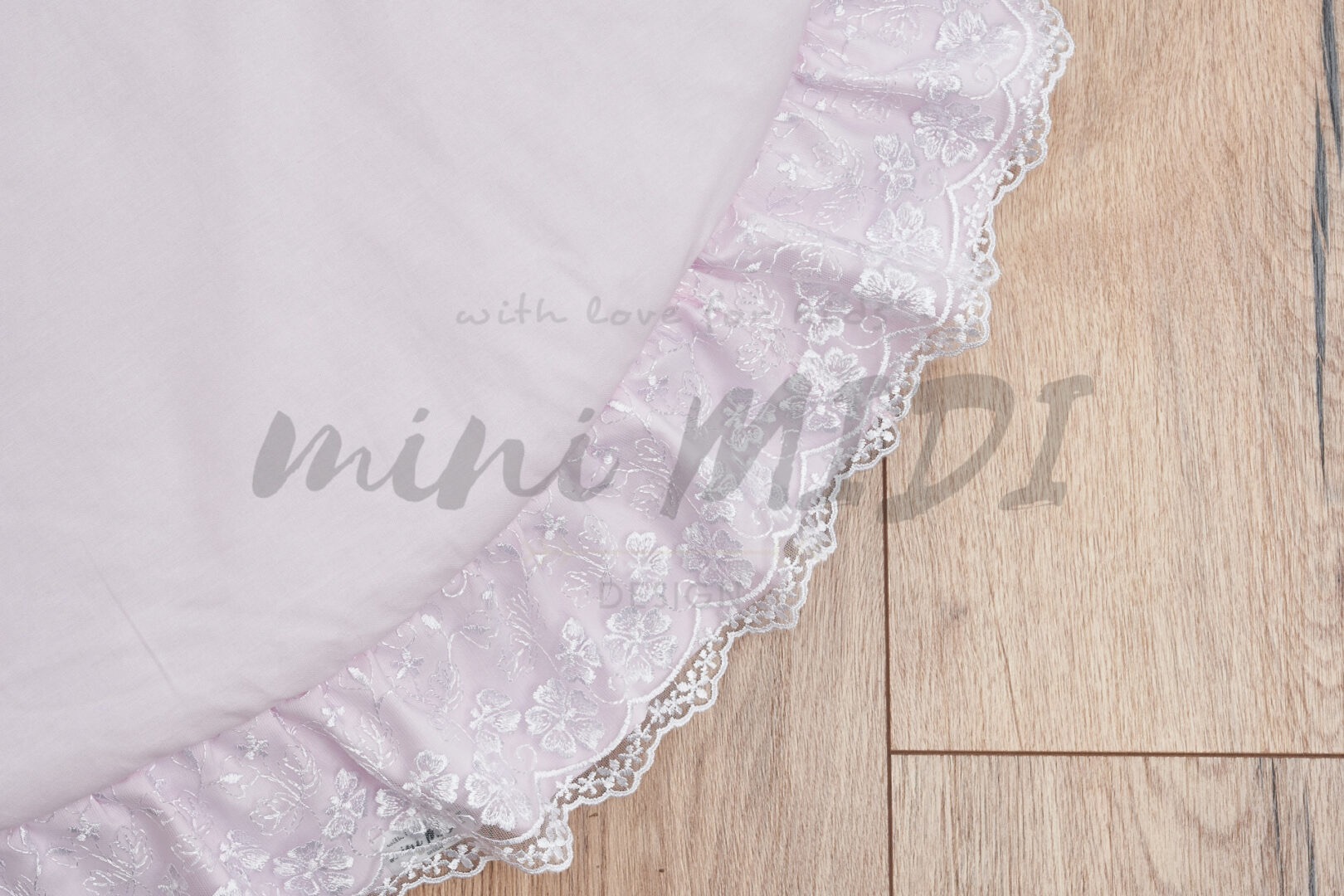 Jasnoróżowa, okrągła poduszka z falbaną obszytą białą koronką na drewnianym parkiecie, z delikatnym logo firmy widocznym na poduszce.