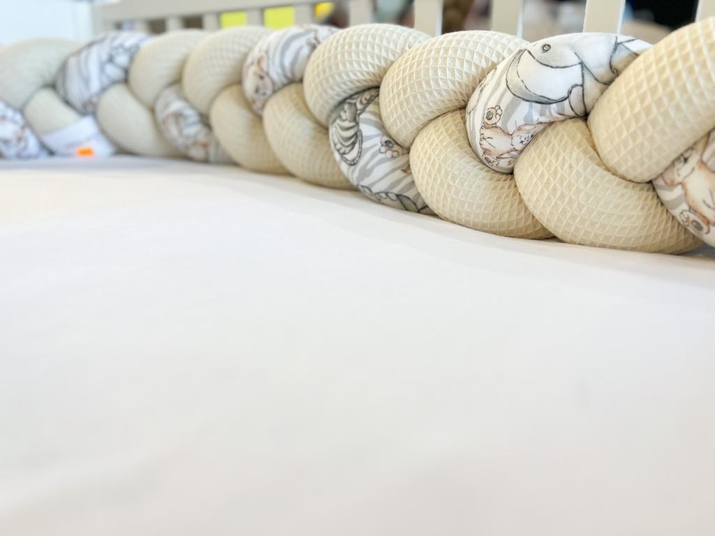 Beżowy, pleciony ochraniacz do łóżeczka dziecięcego z motywem zwierzęcym, ułożony na białym prześcieradle, widoczne szczebelki łóżeczka w tle.