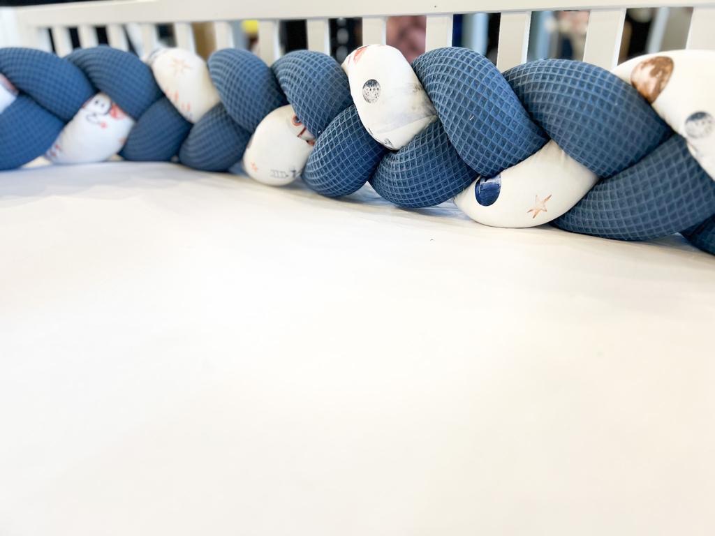 Ochraniacz do łóżeczka dziecięcego w formie warkocza z granatowej i białej dzianiny bawełnianej, leżący na białym prześcieradle w łóżeczku.