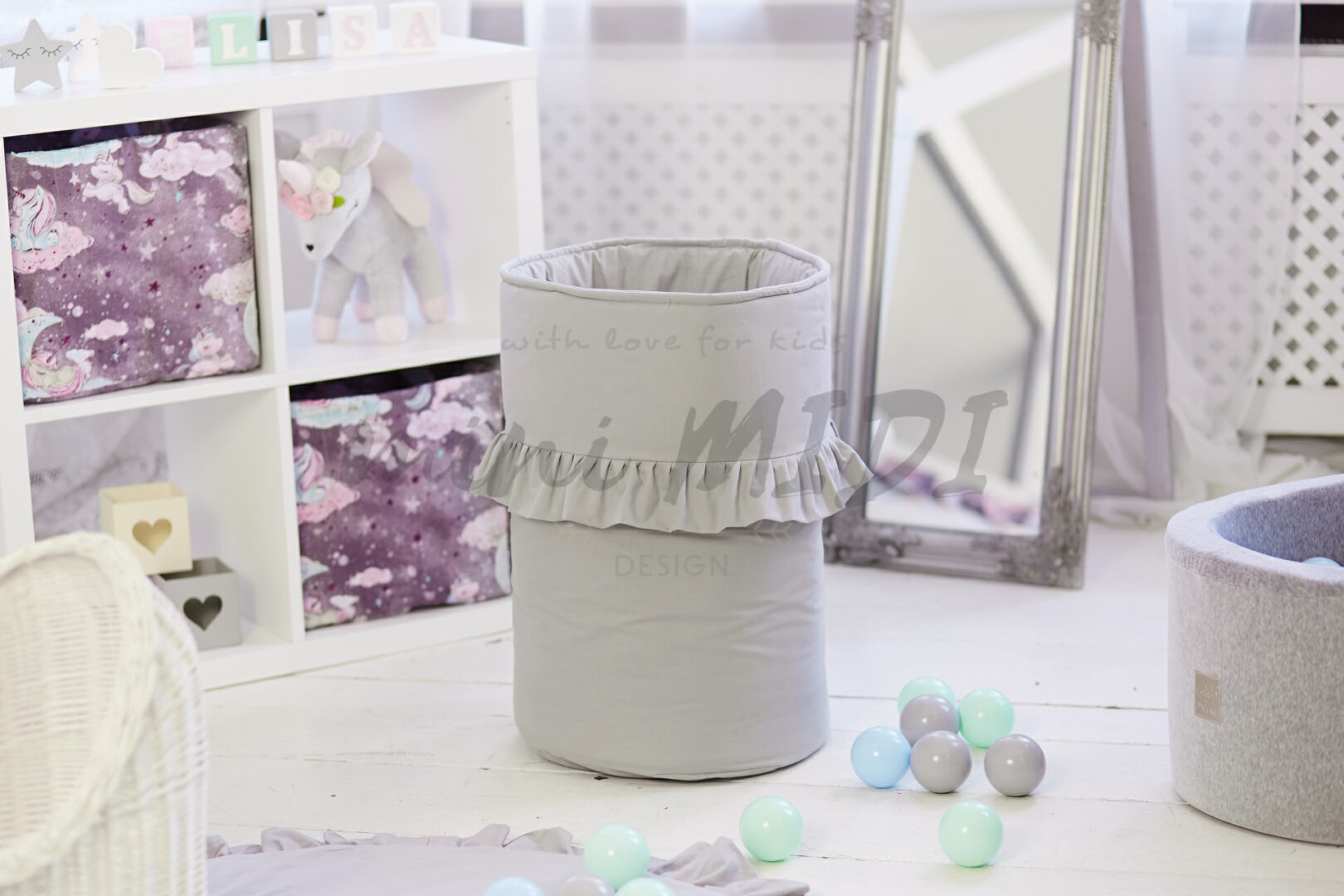 Szary, tekstylny kosz na zabawki z falbanką, ustawiony na białej podłodze pokoju dziecięcego, obok lustra w srebrnej ramie i regału z pudełkami w jednorożce.