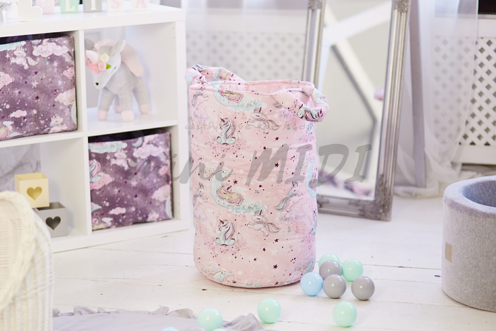 Różowy, materiałowy kosz na zabawki z motywem jednorożców, stojący na białej podłodze w pokoju dziecięcym, obok kolorowe piłeczki i regał z pudełkami.