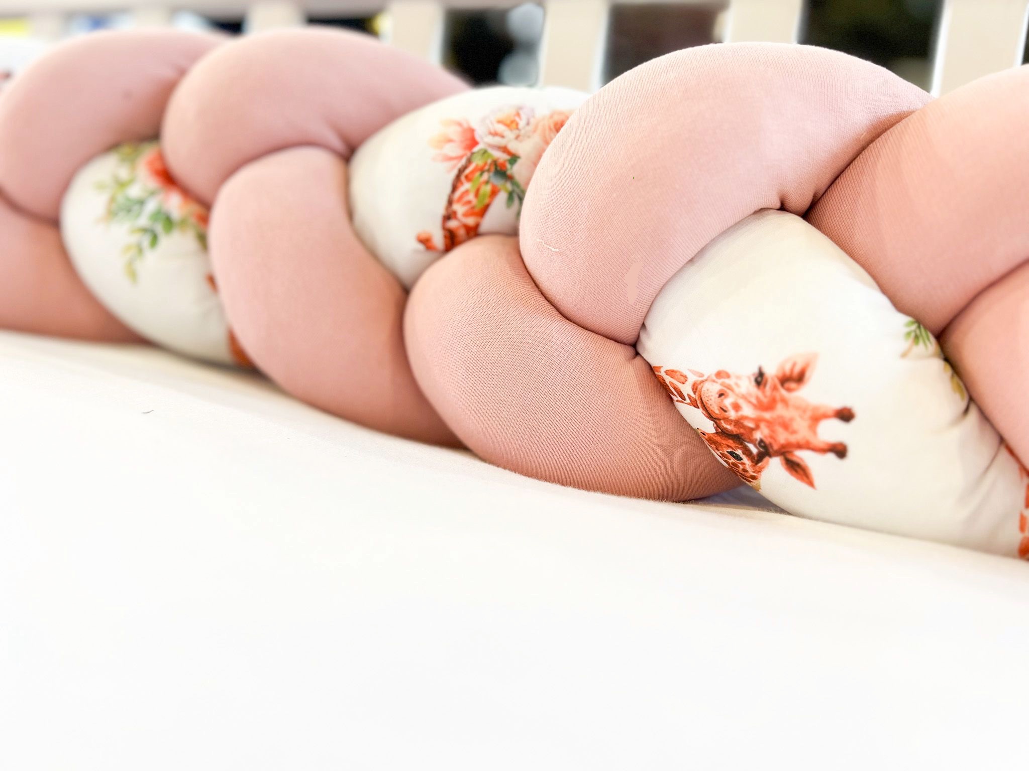 Ochraniacz do łóżeczka dziecięcego w formie warkocza, uszyty z różowej tkaniny i białej w kwiaty i żyrafy, leżący na białym prześcieradle.