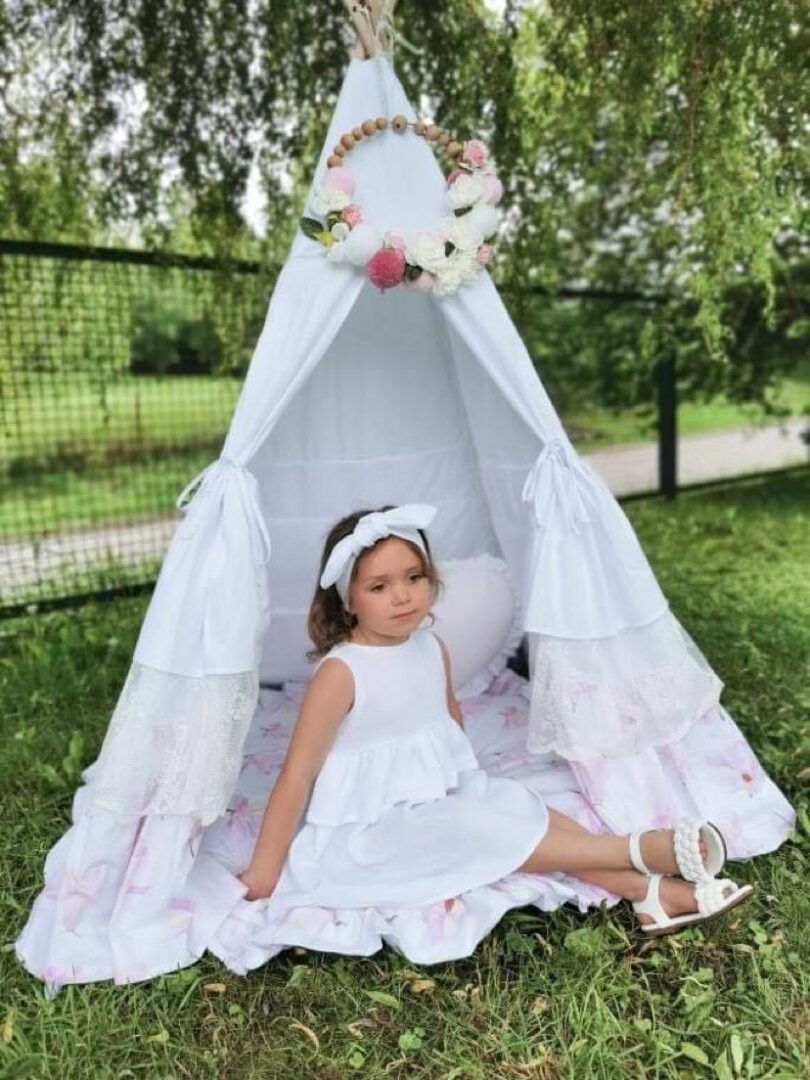 Białe tipi dla dziecka z dekoracją kwiatową, dziewczynka w białej sukience i sandałach siedzi w środku na kwiecistej pościeli, ogród w tle.