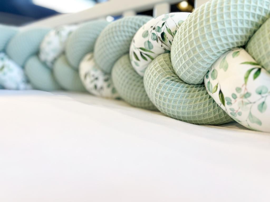 Jasnozielony, pleciony ochraniacz do łóżeczka dziecięcego z wzorem liści eukaliptusa, leżący na białym prześcieradle, widok z bliska.