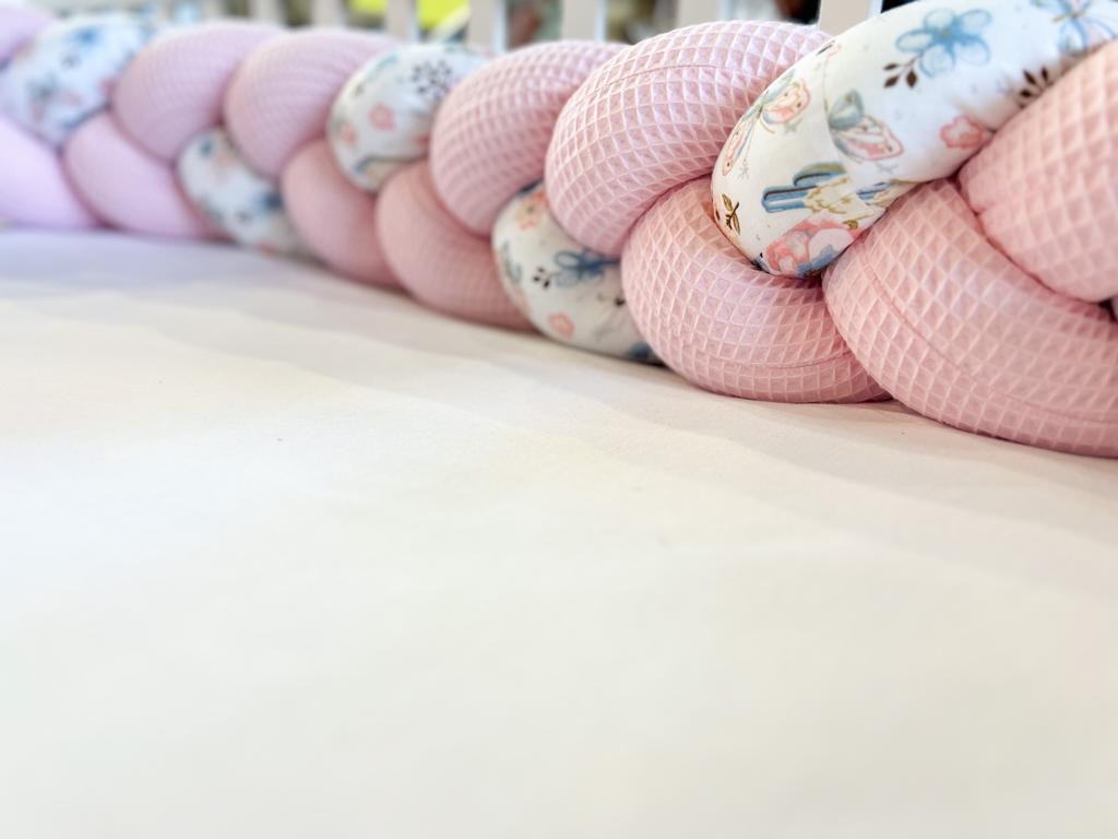 Ręcznie pleciony, różowo-biały ochraniacz do łóżeczka dziecięcego z tkaniny wafelkowej i wzorzystej bawełny, leżący na białym prześcieradle.