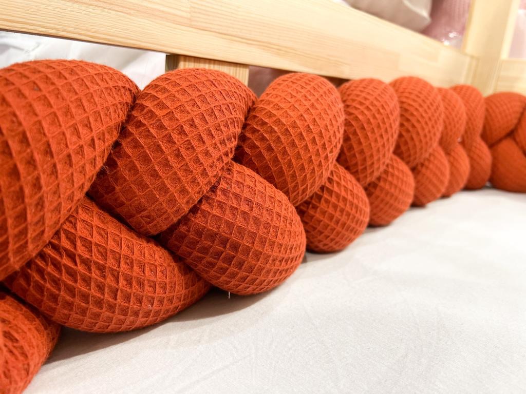 Pomarańczowy, pleciony ochraniacz na szczebelki łóżeczka dziecięcego z tkaniny o strukturze wafla, leżący na białym prześcieradle.