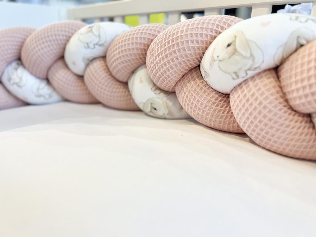 Ochraniacz do łóżeczka dziecięcego w formie warkocza, wykonany z różowej dzianiny wafelkowej i białej tkaniny z motywem króliczków, ułożony na białym prześcieradle.