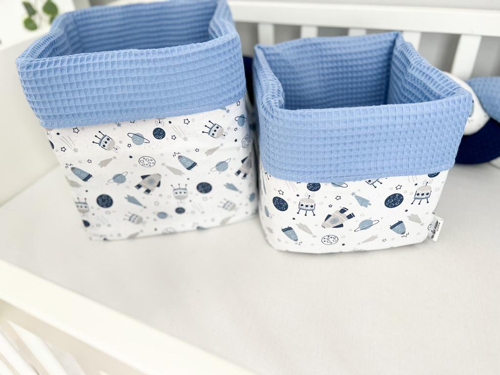 Dwa tekstylne pojemniki na zabawki z motywem kosmicznym, wykończone błękitną waflową tkaniną, stojące na białym łóżeczku dziecięcym.