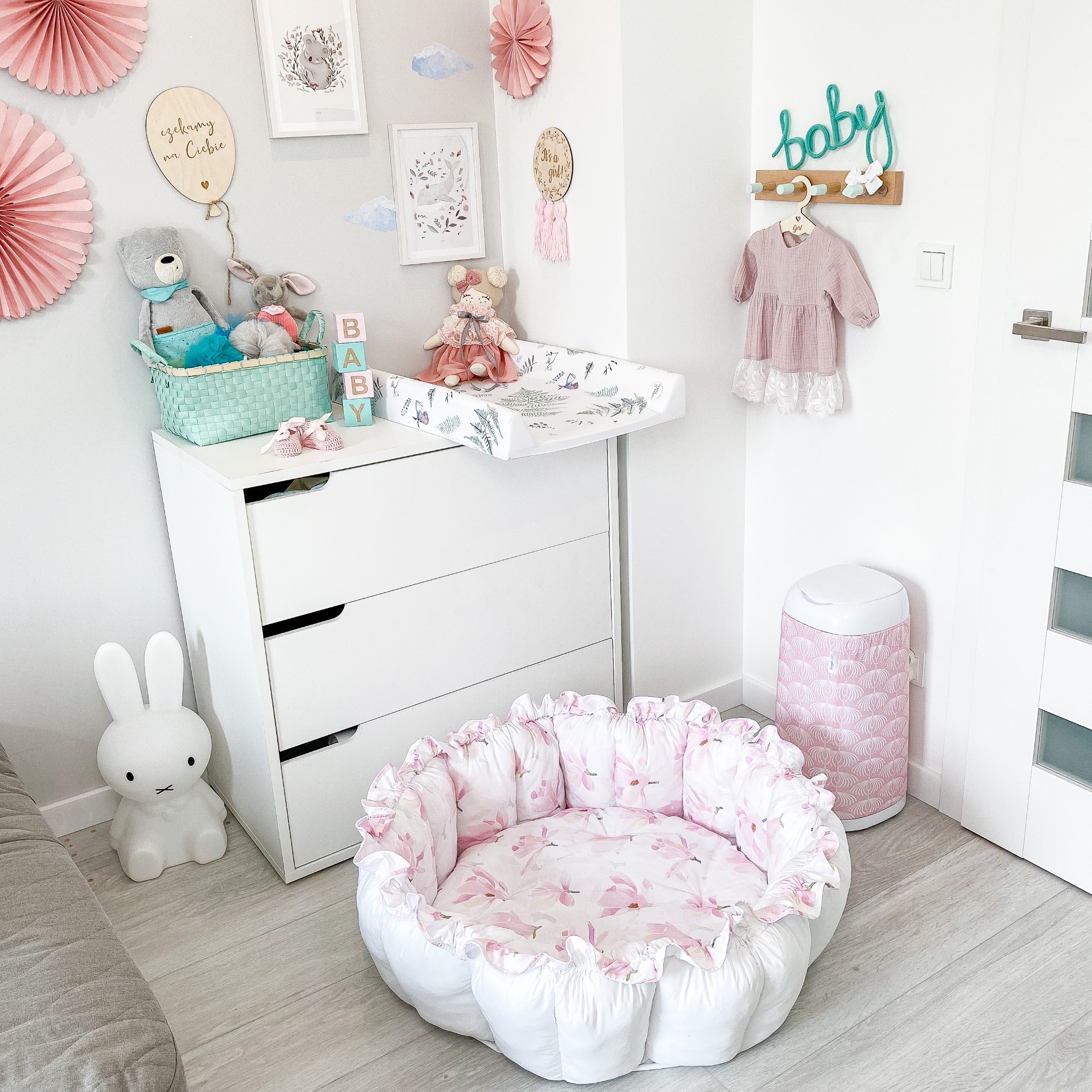 Pokój dziecięcy z białymi meblami, łóżeczkiem w kształcie kwiatu z różowym wzorem, komodą z przewijakiem, dekoracjami ściennymi i lampką nocną w kształcie królika.