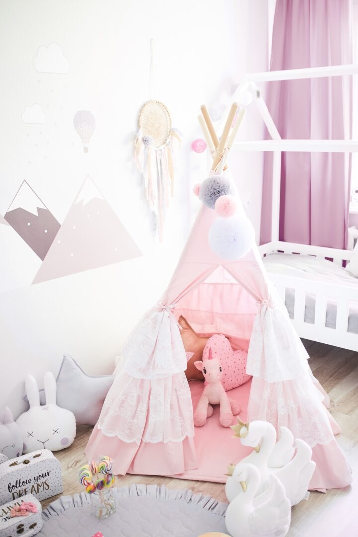 Różowy, koronkowy namiot tipi dla dzieci w pokoju z dekoracjami, wewnątrz poduszki w kształcie serca i jednorożca, obok pluszowe łabędzie i dywanik z falbanką.