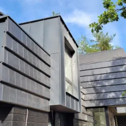 Nowoczesny dom z drewnianą elewacją i dachem pokrytym szarą dachówką ceramiczną oraz blachą na ścianach, widok z dołu na tle drzew i błękitnego nieba.