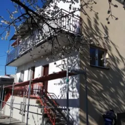 Dwupiętrowy dom z balkonami, na którym widać rusztowania w trakcie ocieplania elewacji, z kwitnącym drzewem w pierwszym planie i pracującym mężczyzną przy ziemi.