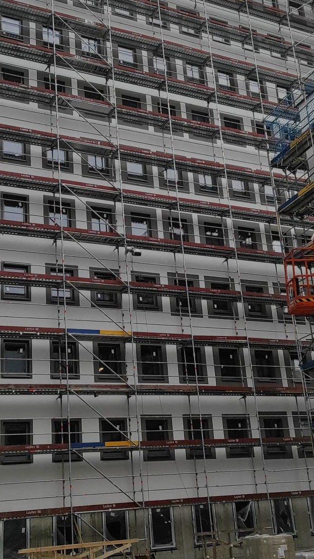 Wielopiętrowy budynek w trakcie prac elewacyjnych, z widocznymi rusztowaniami na całej wysokości fasady i podnośnikiem koszowym po prawej stronie.