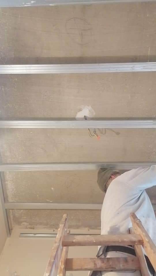 Prace przygotowawcze do montażu sufitu podwieszanego: widoczne metalowe profile na tle surowego sufitu, na którym widać szkic ołówkiem, oraz fragment drabiny i sylwetka pracownika w białym ubraniu.