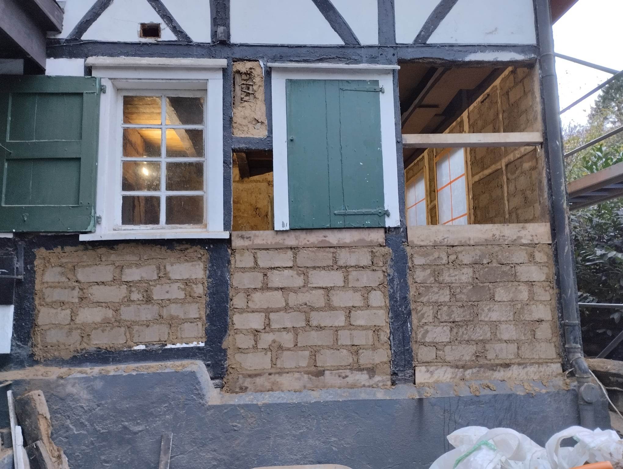 Fragment elewacji domu szachulcowego w trakcie prac murarskich, widoczne cegły i zaprawa gliniana, jedno okno z otwartą zieloną okiennicą, drugie z częściowo otwartą, trzecie bez okna, z widocznym...