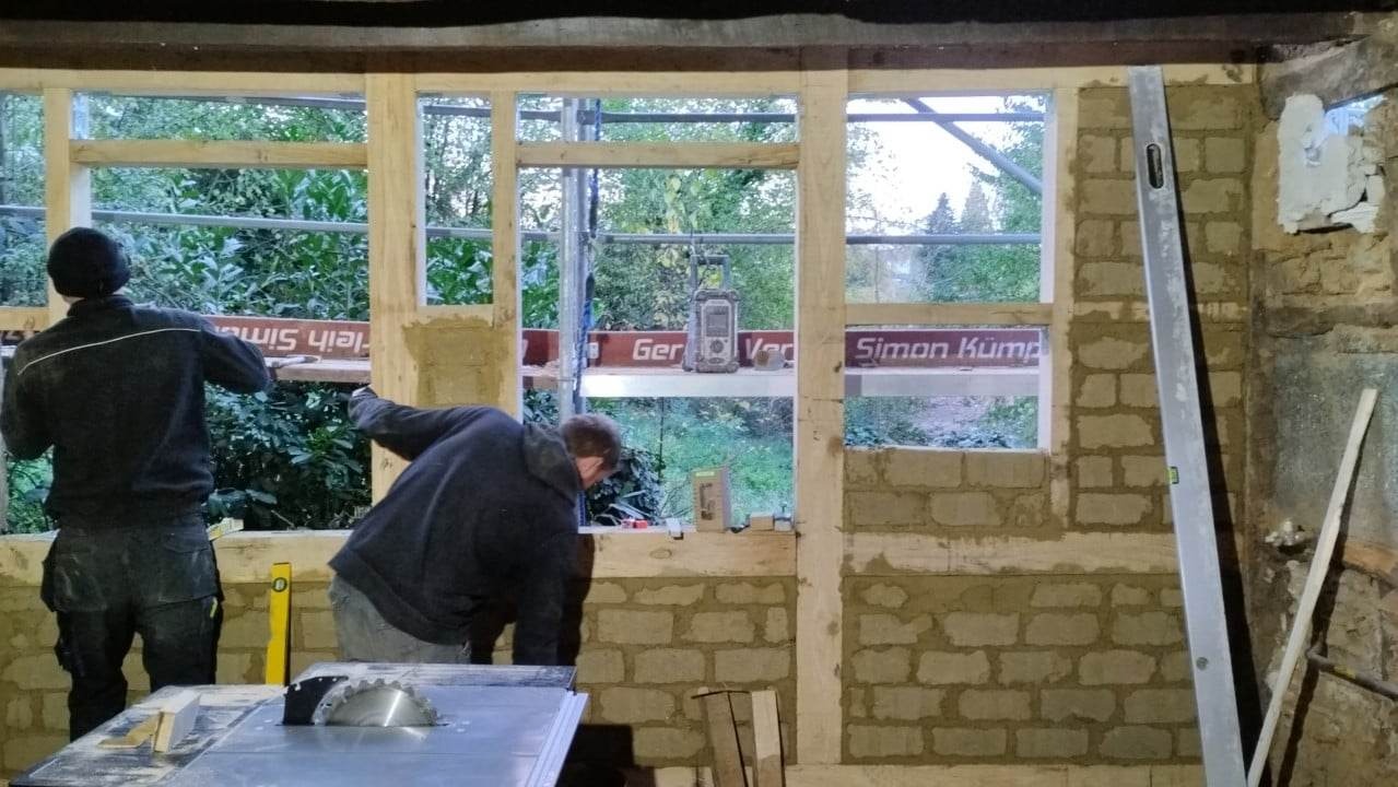 Wnętrze budynku w trakcie murowania ścian z bloczków, widoczni dwaj mężczyźni przy pracy, piła stołowa na pierwszym planie, poziomica oparta o ścianę, okno z widokiem na zieleń.