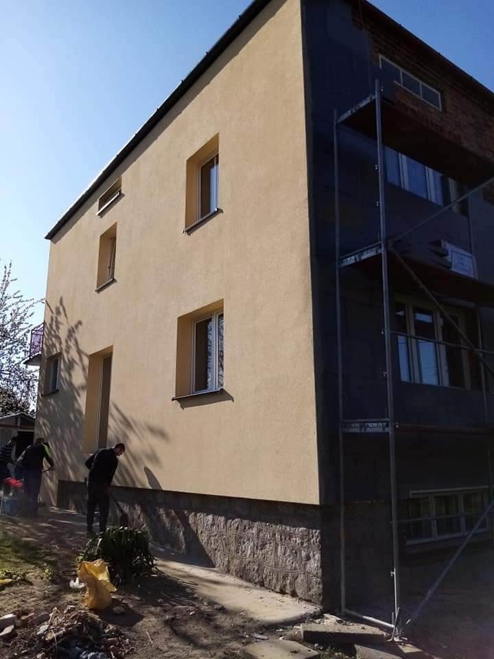 Dwupiętrowy budynek w trakcie renowacji elewacji; jedna ściana w kolorze piaskowym, druga pokryta ciemną warstwą izolacyjną i rusztowaniem; widoczni pracownicy przy dolnej części budynku.