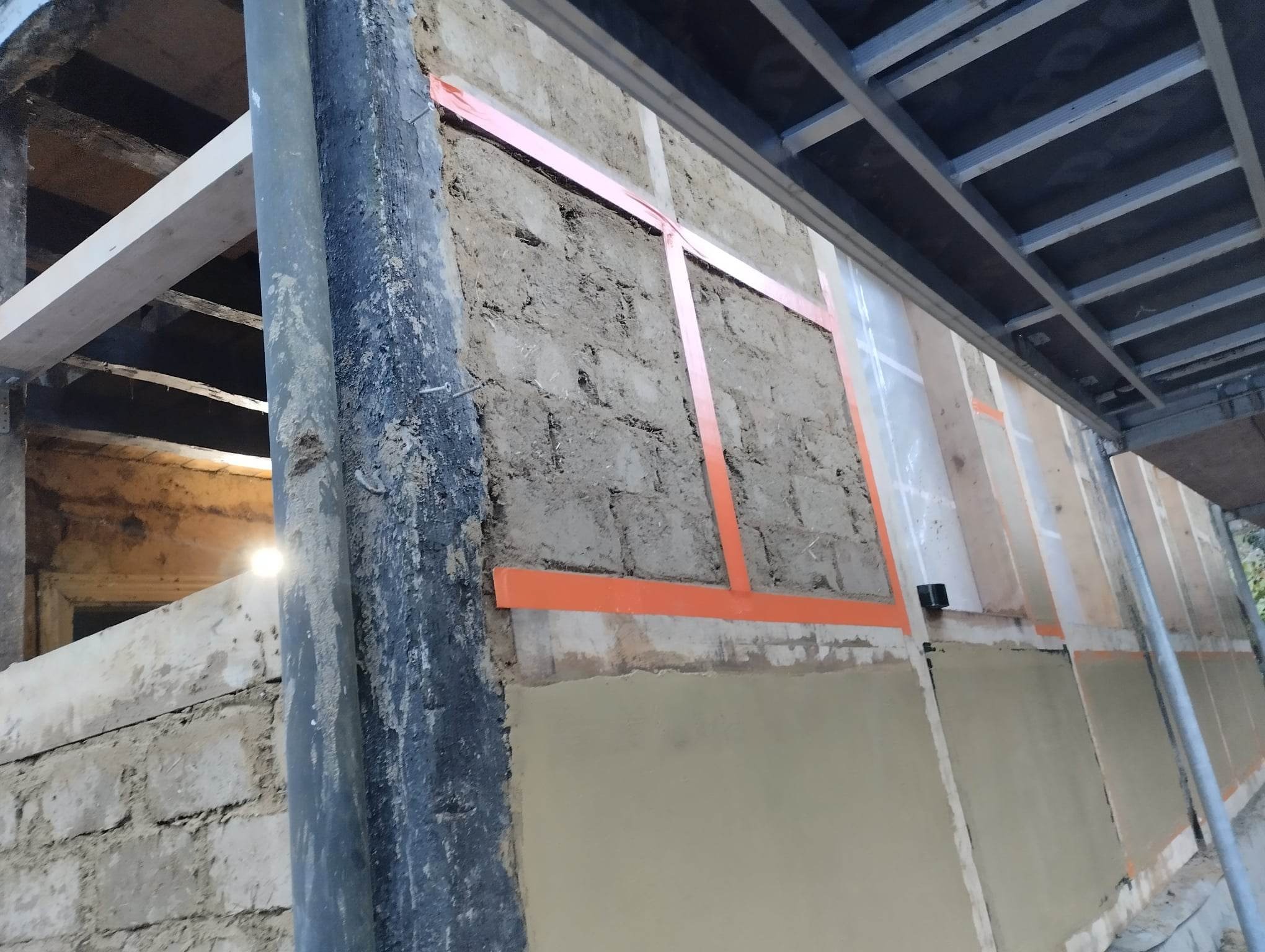 Ściana zewnętrzna budynku w trakcie prac remontowych, widoczne fragmenty starej elewacji z cegły i zaprawy, oraz nowe warstwy tynku i elementy konstrukcyjne rusztowania.