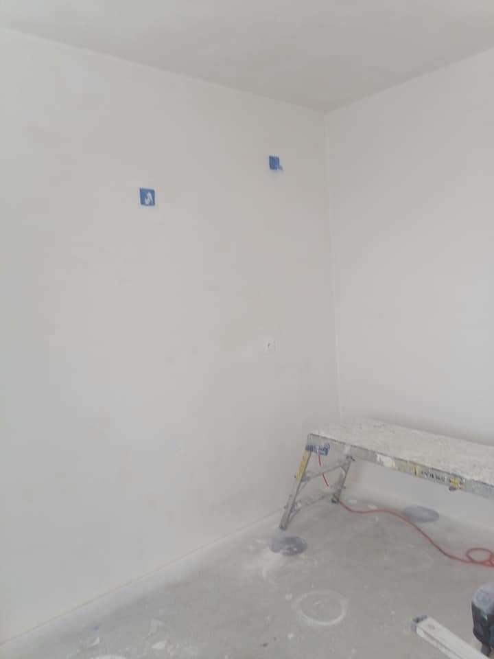 Wnętrze pokoju po szpachlowaniu ścian, widoczne niebieskie taśmy zabezpieczające, drabina malarska i ślady gładzi na podłodze.