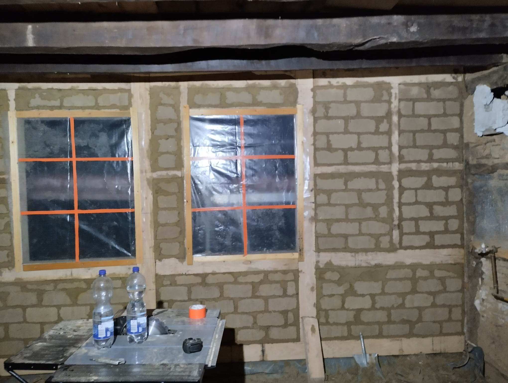 Wnętrze w trakcie murowania ścian z bloczków, widoczne dwa okna zabezpieczone folią i taśmą, na pierwszym planie stół roboczy z butelkami wody i narzędziami.