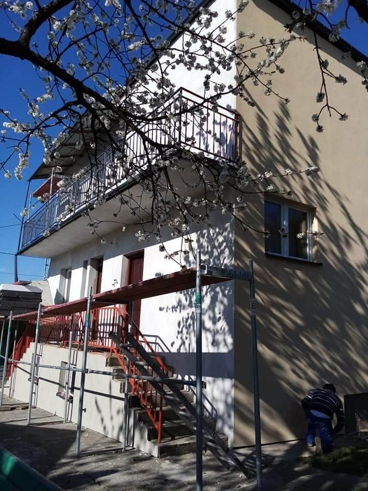 Dwupiętrowy dom z balkonami, na którym widać rusztowania w trakcie ocieplania elewacji, z kwitnącym drzewem w pierwszym planie i pracującym mężczyzną przy ziemi.