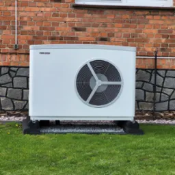 Biała jednostka zewnętrzna z logo Stiebel Eltron na tle ceglanego domu z oknem i zielonym trawnikiem.