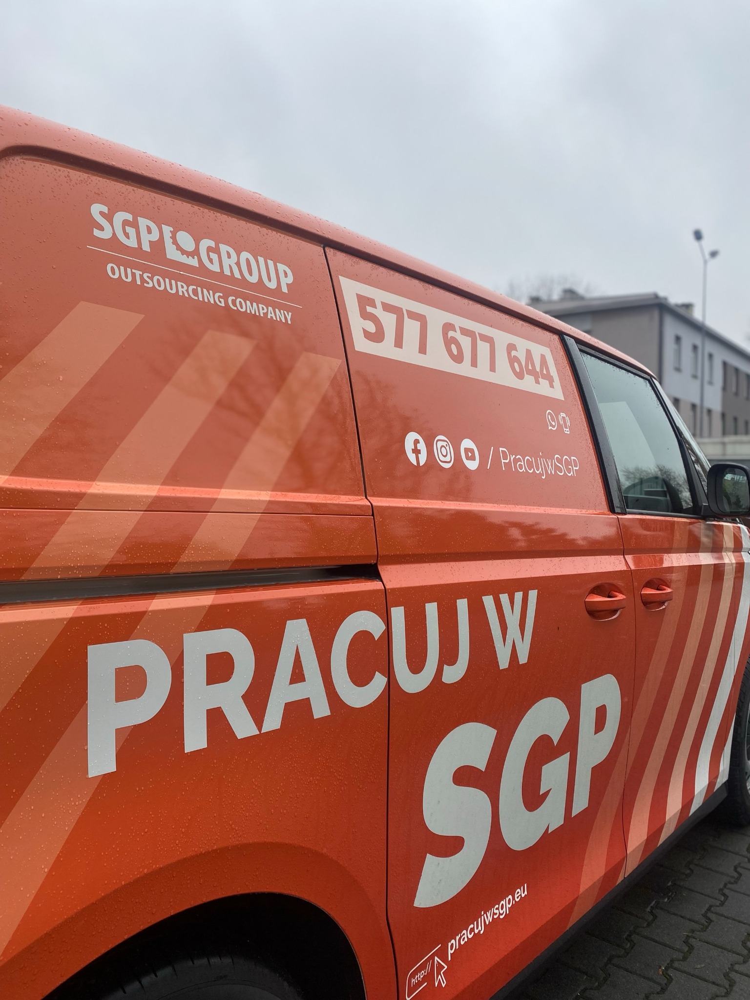 Pomarańczowy samochód dostawczy z logo SGPE Group Outsourcing Company, numerem telefonu i odnośnikami do mediów społecznościowych na tle miejskim w pochmurny dzień.