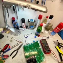 Stół warsztatowy z rozłożoną płytą główną z wentylatorem, multimetrem, narzędziami i środkami czyszczącymi, oświetlony lampą warsztatową, sugerujący naprawę sprzętu elektronicznego.