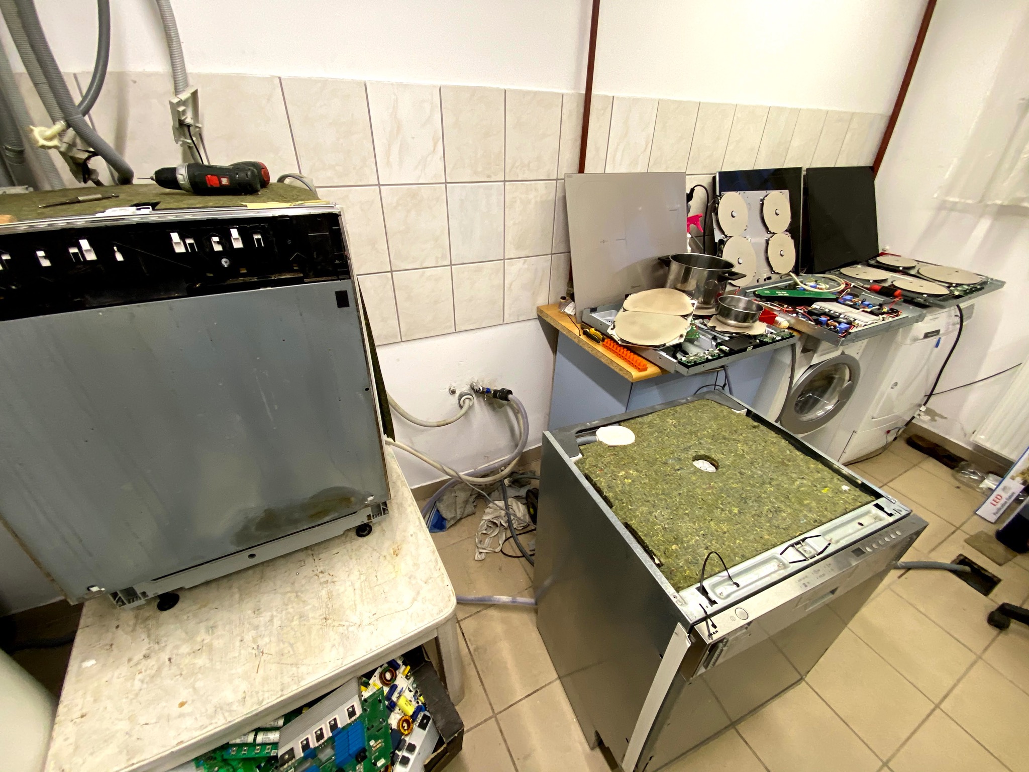 Dwie zmywarki w trakcie demontażu w pomieszczeniu z białymi kafelkami, jedna z odkręconym blatem leży na stole, druga stoi na podłodze z widocznymi obwodami elektronicznymi na podłodze i pralką w tle.