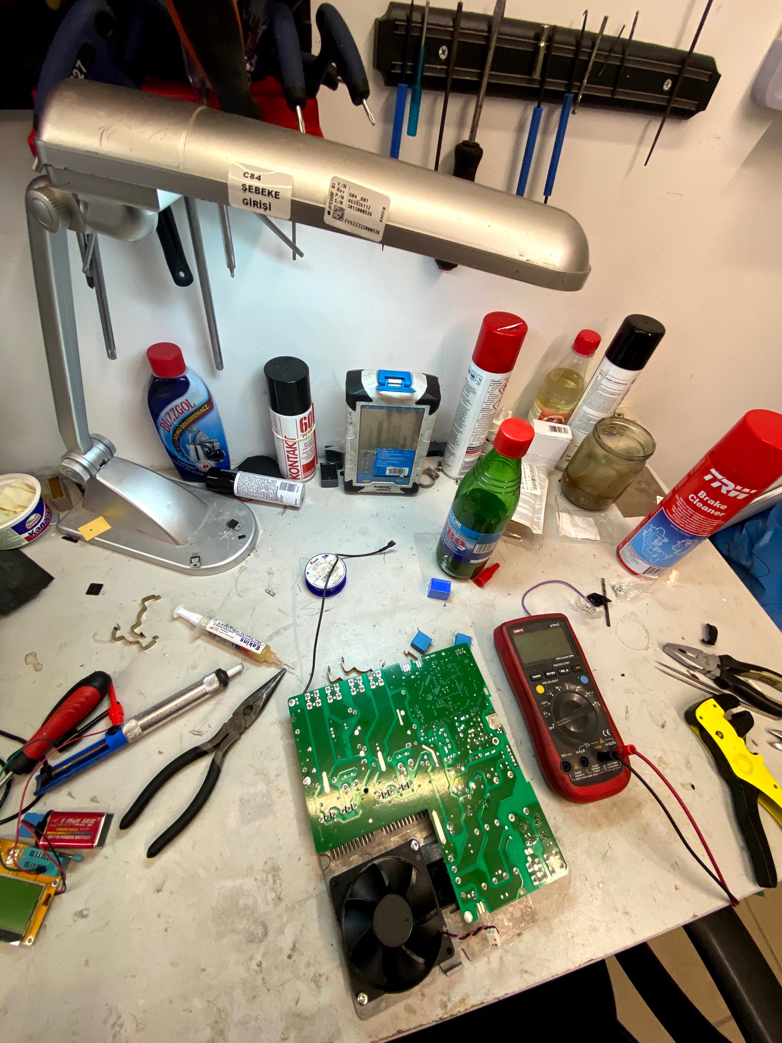 Stół warsztatowy z rozłożoną płytą główną z wentylatorem, multimetrem, narzędziami i środkami czyszczącymi, oświetlony lampą warsztatową, sugerujący naprawę sprzętu elektronicznego.